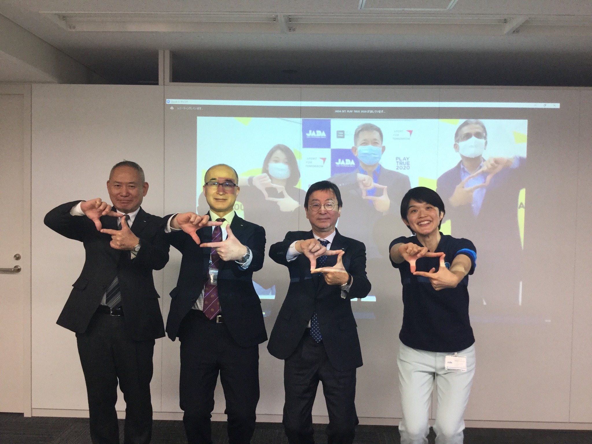 Four people in formal attire stand together making finger frame gestures in front of a JADA Play True 2020 presentation screen.